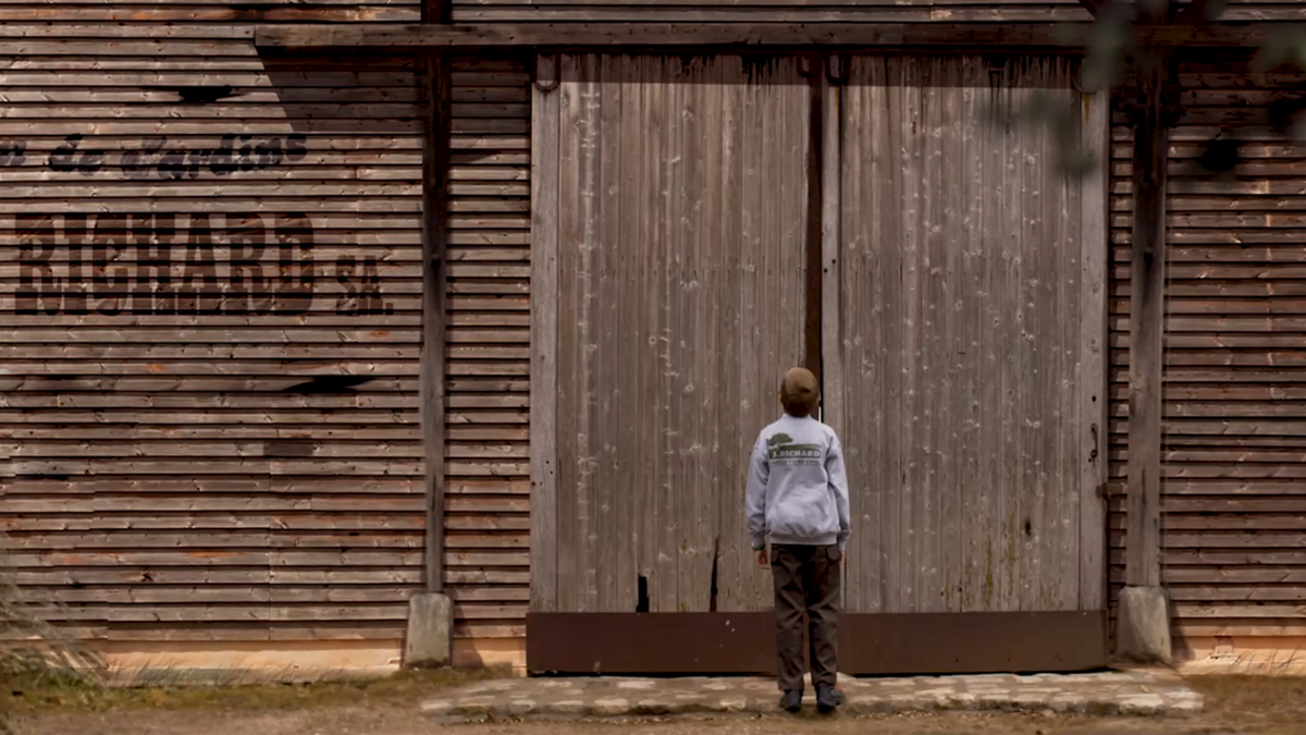 Un enfant en sweat J.Richard face aux grandes portes en bois d'un hangar