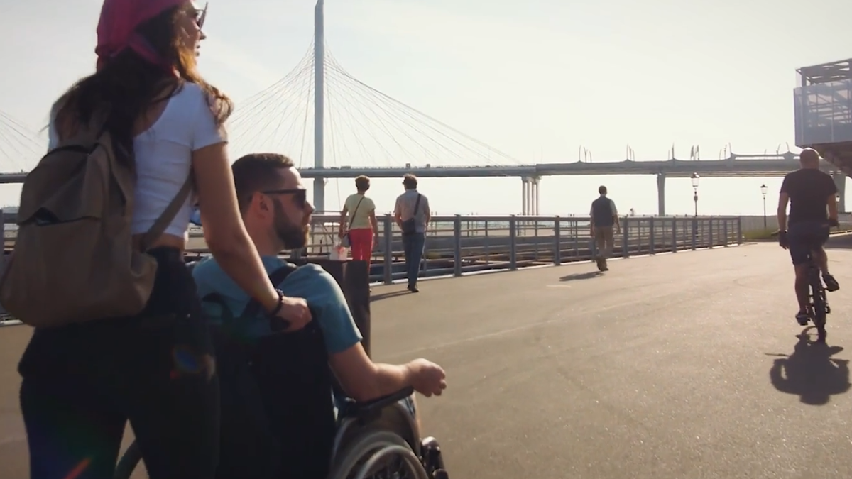 Un homme en fauteuil roulant accompagné d'une femme sur une promenade avec un pont moderne en arrière-plan