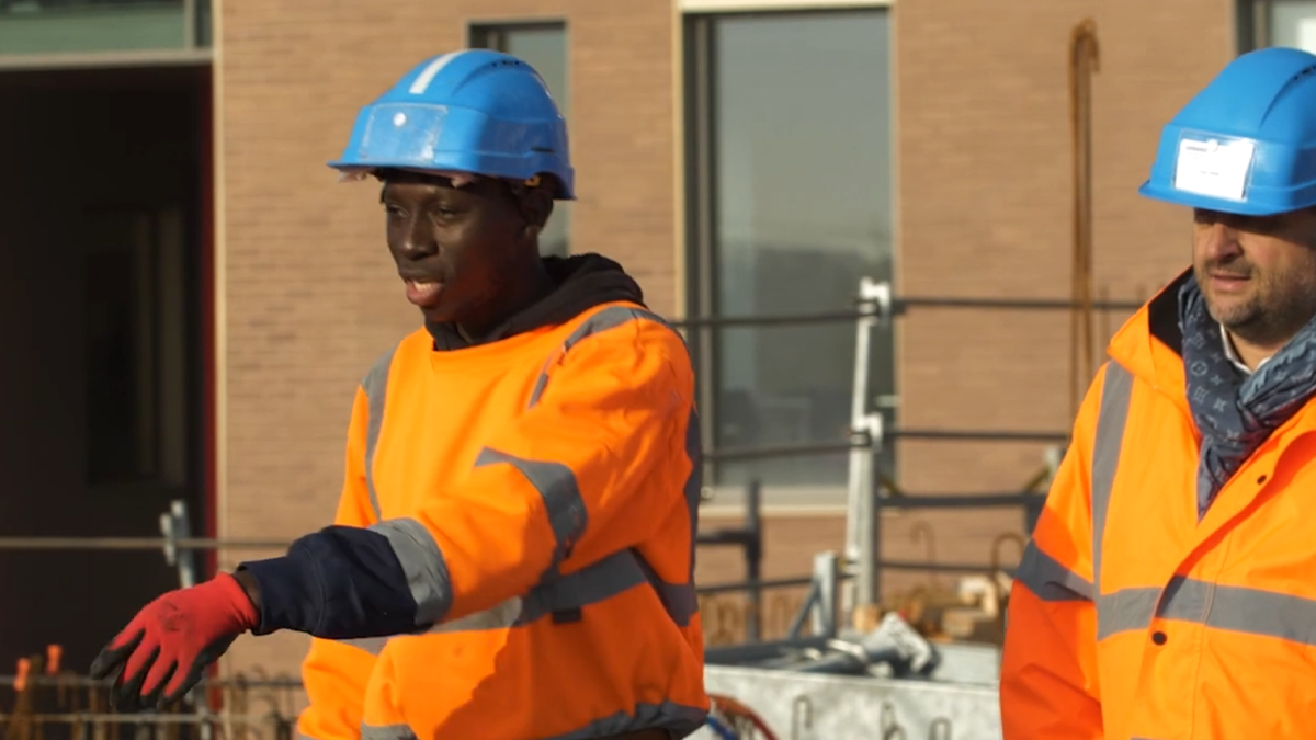 Ouvrier du BTP en gilet orange et casque bleu s'exprimant sur un chantier