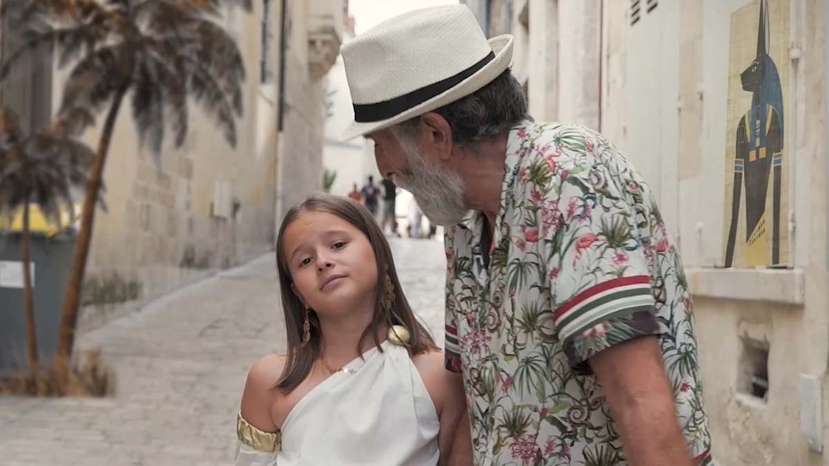 Une fillette en tenue antique blanche accompagnée d'un homme âgé, dans une ruelle méditerranéenne
