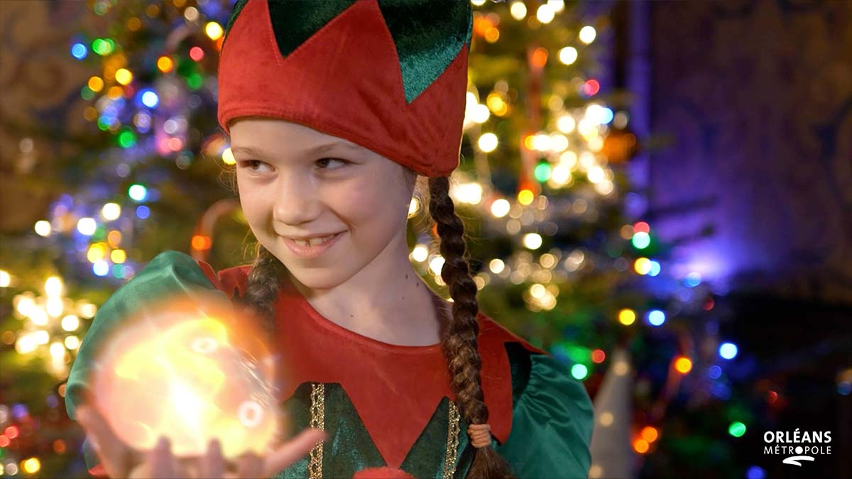 Une fillette déguisée en lutin tenant une boule lumineuse devant un sapin de Noël illuminé